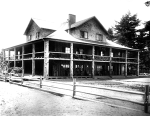 A large wooden house with a peaked roof and wrap around porch on the first and second story. The porch columns are rough hewn logs. Wooden letters on the porch railings say The Stockade. 